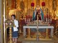 Una mujer se encuentra en el altar de una iglesia, frente a un altar dorado con una imagen de la Virgen María. La decoración es elaborada y dorada, reflejando un ambiente religioso serio.