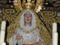 Imagen de una Virgen María vestida con un manto blanco y una corona dorada, rodeada de estrellas y luces. Detalles dorados en el fondo.