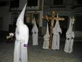 Procesión de la Semana Santa con nazarenos en túnicas blancas y capas, portando cruces y faroles en una calle adoquinada de noche.