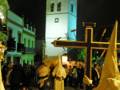 Procesión de Semana Santa con penitentes portando una cruz en la noche, con una iglesia iluminada al fondo.
