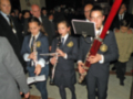 Un grupo de niños en uniforme escolar toca instrumentos musicales, incluyendo clarinetes y flautas. La imagen muestra a los niños concentrados en su interpretación, con adultos observando desde atrás.