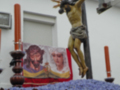 Imagen de un crucificado en la cruz, con una imagen de Jesús y una figura masculina en el fondo. La escena se encuentra en un entorno urbano con un edificio blanco y una ventana visible. La imagen está iluminada por la luz del día, creando un contraste entre las sombras y los detalles de la escena.
