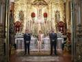 Se observa a tres personas vestidas formalmente, posando frente a un altar de una iglesia. El altar está ricamente decorado con estatuas, flores y candelabros dorados. La escena transmite una atmósfera de solemnidad y respeto religioso.