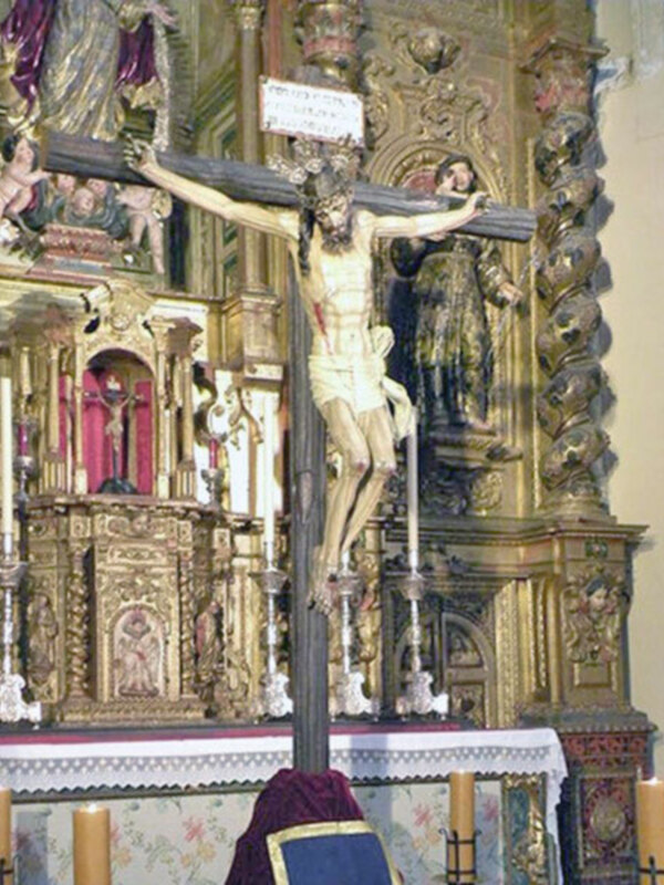 Imagen de un altar con estatuas religiosas y velas encendidas, enfocándose en la figura central de un Cristo crucificado. El estilo barroco y dorado destaca en el fondo, con detalles escultóricos elaborados.