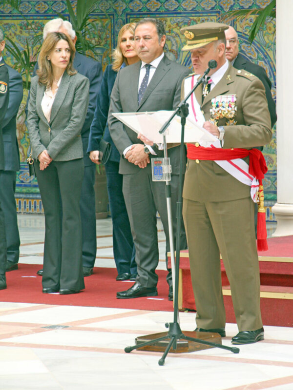 Una imagen de una ceremonia formal en un edificio con decoración tradicional, donde varios hombres y mujeres vestidos de manera elegante se encuentran en un escenario rojo. Algunos están hablando al micrófono, mientras otros observan la escena. La decoración del edificio es impresionante con detalles en azul y verde, lo que sugiere un ambiente de respeto y solemnidad.