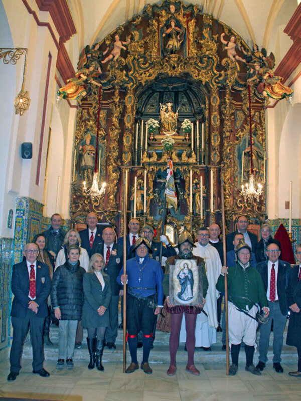 Una imagen de una ceremonia en un templo o iglesia, con varias personas vestidas formalmente. En el fondo, se puede ver un altar elaborado y una araña de luces colgantes. La decoración del interior es elegante, con detalles dorados y azules.