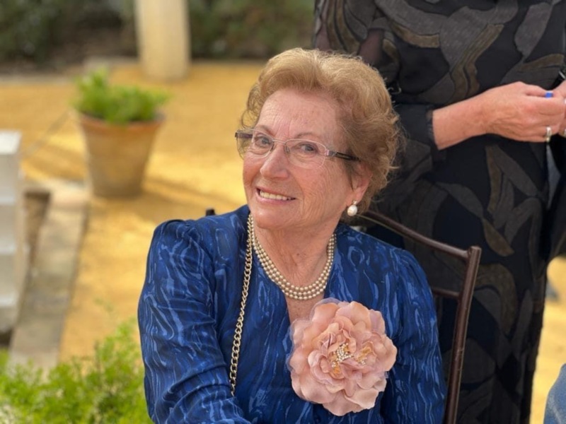 Una anciana sonriente con cabello canoso y una blusa azul, posa frente a un jardín. La imagen está tomada en una terraza con baldosas y plantas verdes al fondo.