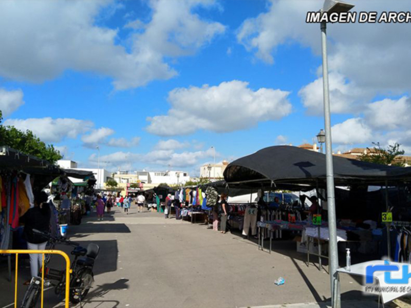 Mercado de abastos en una ciudad con edificios y árboles al fondo. El cielo está lleno de nubes blancas y el suelo es de asfalto. Hay varios estand con ropa colgada y un poste de luz en la parte derecha.