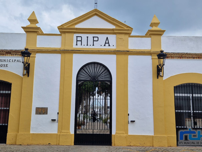 Fachada de un edificio blanco y amarillo con la inscripción "R.I.P.A." en el techo. La entrada tiene una puerta metálica y ventanas con persianas. El edificio parece ser un lugar público o institucional.