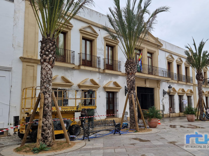 Edificio blanco con balcones y palmeras, en un entorno urbano con una máquina de construcción amarilla.