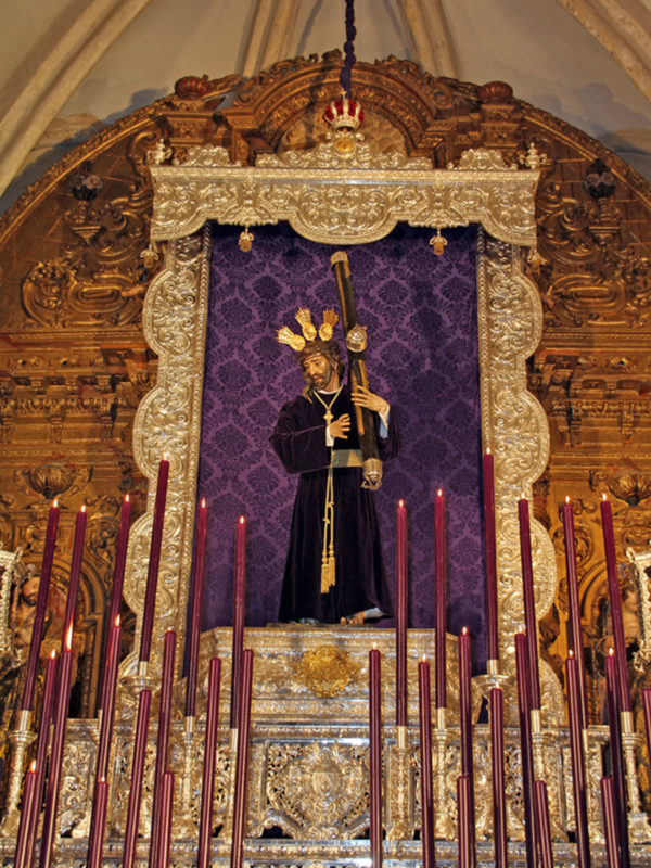 Un altar de madera tallada con un estatua de un sacerdote en el centro, rodeado por candelabros rojos y una cortina de terciopelo morado. La arquitectura del altar es elaborada con detalles dorados y esculturas de ángeles.
