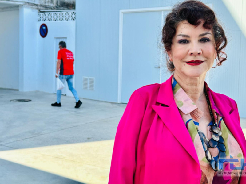 A una mujer con un vestido rosa brillante y un collar de perlas. En el fondo, se ve a otra persona caminando con una bolsa blanca. La escena parece ser en un edificio moderno con paredes blancas y una puerta abierta.