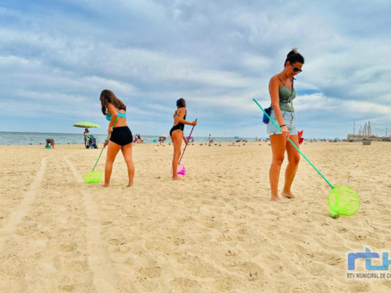 Tres personas en la playa, dos niñas y una adulta, jugando con redes de pesca. El cielo es azul con algunas nubes. La arena es clara y brillante, y hay otros bañistas en el fondo.