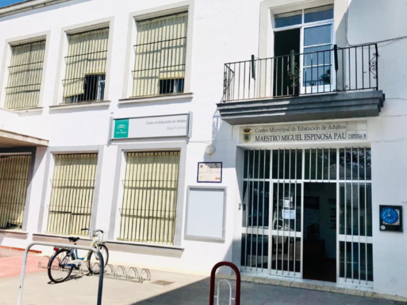Un edificio blanco con ventanas y un balcón. En la entrada, hay una señal de aparcamiento para bicicletas y un cartel con información. La imagen parece ser de una oficina o centro administrativo.
