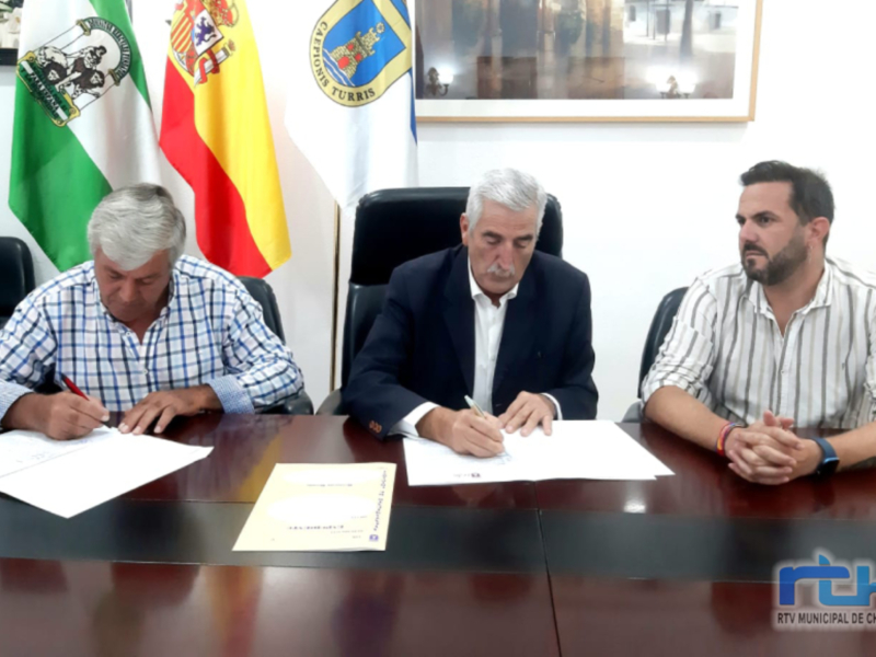 Tres personas firmando documentos en una oficina, con banderas y un escudo de armas en el fondo.
