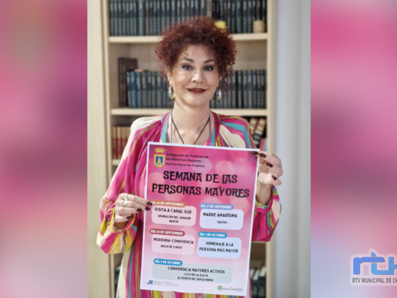 Una mujer con cabello rojo muestra un cartel informativo sobre la 'Semana de las Personas Mayores' en el Ayuntamiento de Chipiona, con actividades como visitas a la televisión y merienda con convivencia.