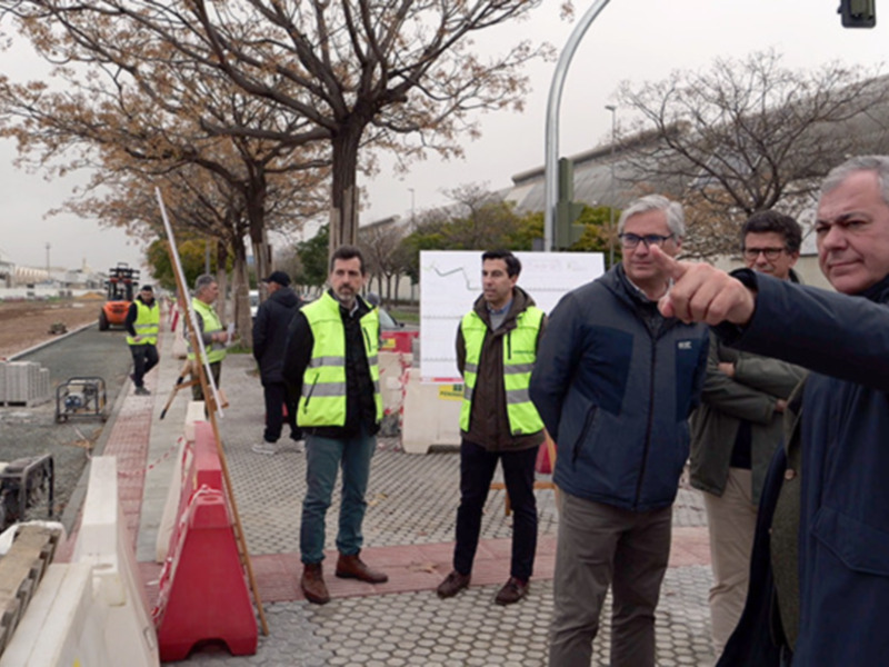 Un grupo de personas con chalecos amarillos y cascos trabajando en una obra. Un hombre apunta hacia algo con su dedo índice, mientras otros observan la construcción en un entorno urbano con árboles secos y una estructura de hormigón.