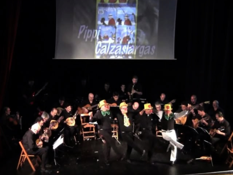 Concierto de música clásica en un escenario con un grupo de músicos y bailarines. La pantalla proyecta imágenes relacionadas con la obra musical "Pippi Galgaskas".