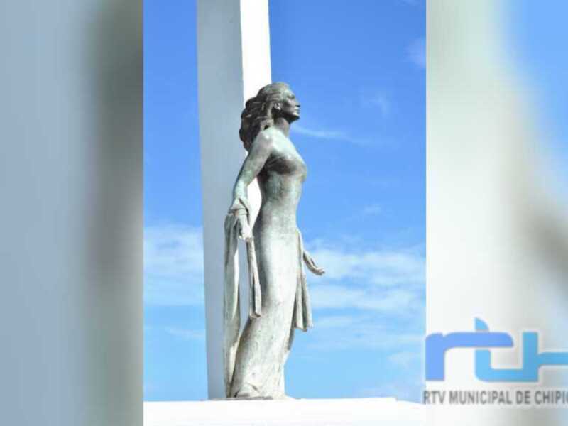 Estatua de una mujer en un monumento, con el cielo azul y la insignia de RTVMunicipal de Chipiona en la esquina inferior derecha.