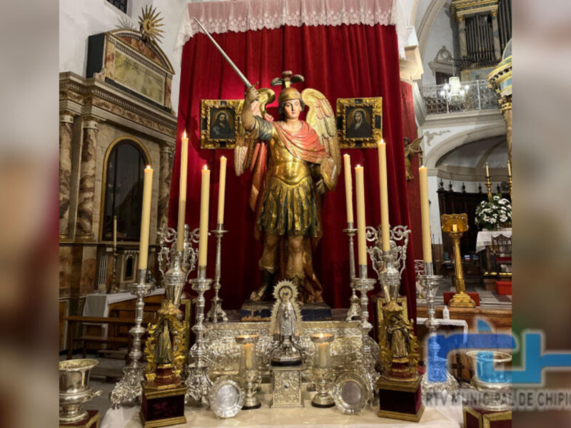 Imagen de un altar con una estatua dorada de San Miguel Arcángel, rodeado por velas y objetos religiosos. El fondo es rojo con un manto y estatuas de ángeles. La imagen está en una iglesia con detalles arquitectónicos y un órgano.