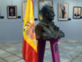Una bandera española ondea en un pedestal rojo, con un busto de bronce y varias pinturas en el fondo. La sala tiene suelo de mármol y paredes grises.