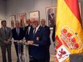 Un hombre en traje negro se encuentra detrás de un micrófono, frente a una bandera española y varios retratos. La escena parece ser un evento formal o ceremonial en una sala con iluminación de techo.