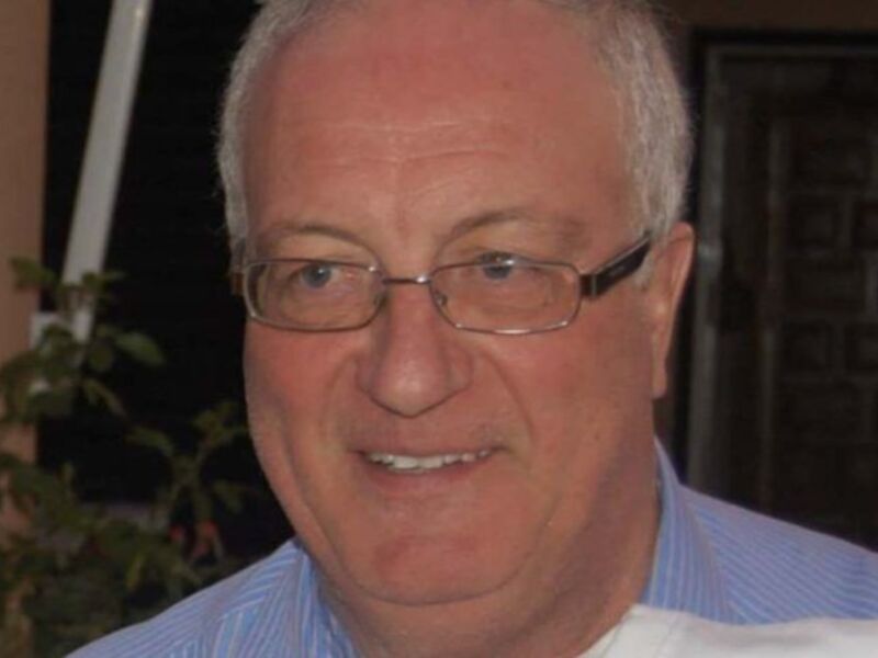 El hombre está sonriendo y lleva anteojos. La imagen parece ser de un entorno doméstico, con una puerta y plantas en el fondo.