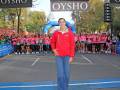 Una mujer en una chaqueta roja y jeans está frente a un grupo de corredores en una carrera. El fondo muestra un arco azul y los nombres "OYSHO" en letras blancas.