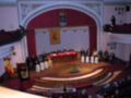 Una sala de conferencias con una mesa central donde se sientan varios hombres. En la parte superior, hay un escudo y una bandera española. La audiencia está sentada en filas de sillas rojas.