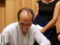 Un hombre mayor firmando un documento mientras una mujer observa en el fondo.