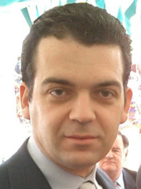 Un hombre con cabello oscuro y peinado hacia atrás, lleva un traje formal. La imagen parece ser de una celebración o evento público debido a la presencia de banderas y decoraciones al fondo.