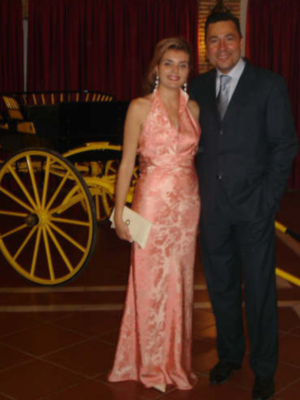 Una mujer vestida con un vestido rosa de encaje largo posa junto a una persona vestida con traje y corbata, frente a un fondo de cortinas rojas y una bicicleta antigua.
