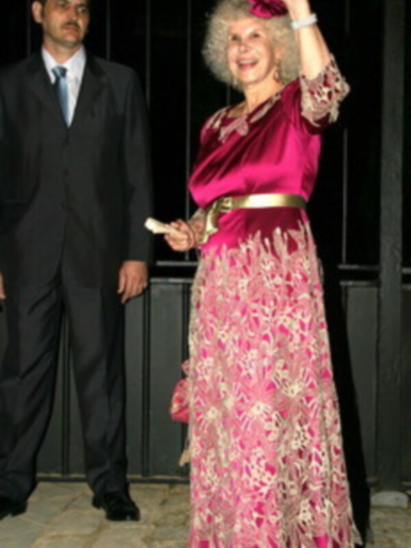 Una mujer elegante con vestido rosa y flores, saluda con la mano mientras está acompañada por otra persona en traje.