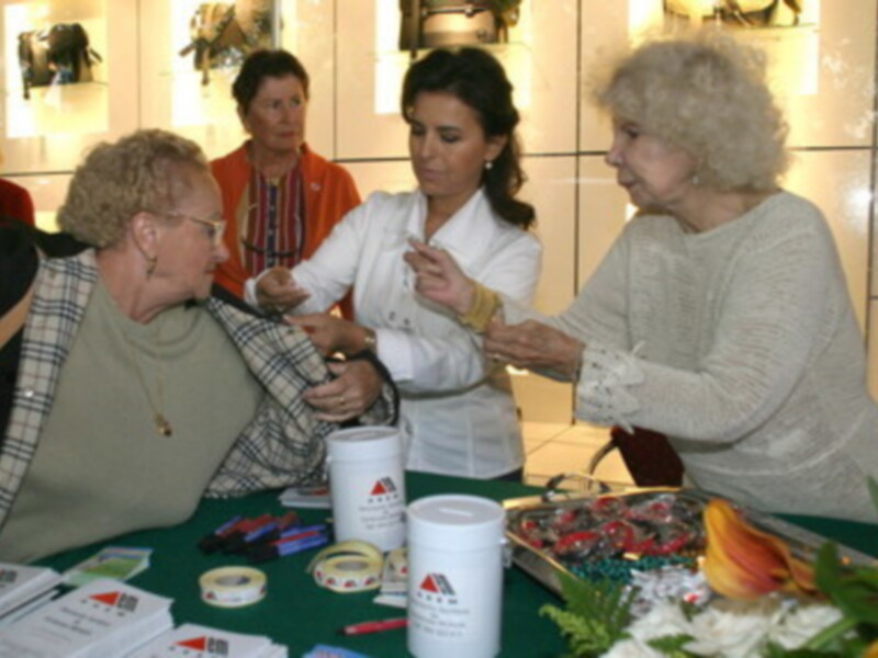 Una mujer en blanco está ofreciendo un regalo a una anciana, mientras que otra mujer observa la escena. La imagen muestra un ambiente de fiesta o celebración, con una mesa cubierta y regalos en el centro.