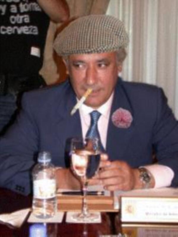 Un hombre con una camisa azul, corbata y sombrero de copa. Está sentado en un escritorio con una botella de agua, un vaso vacío y un libro. Parece estar en una reunión o conferencia formal.