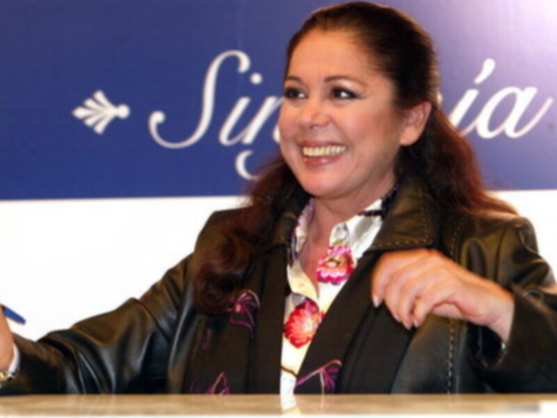 Una mujer sonriendo y hablando en una conferencia de prensa con un fondo azul y blanco.