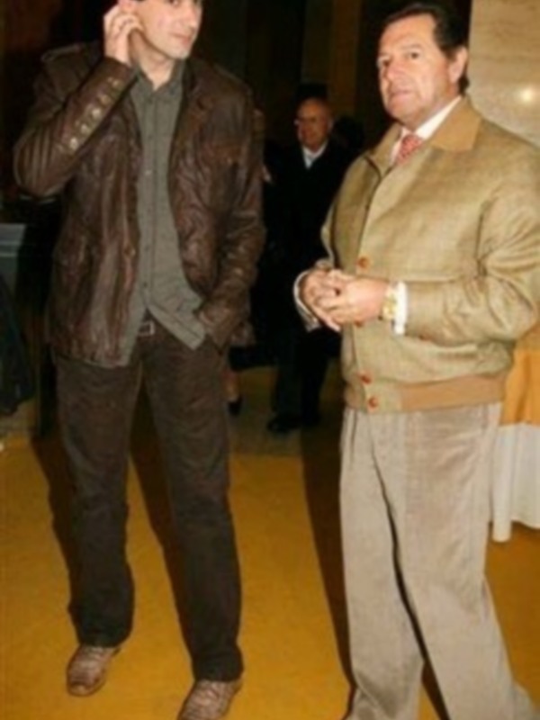 Dos hombres en una sala de eventos, uno hablando por teléfono y el otro en pose de conversación, ambos vestidos formalmente.