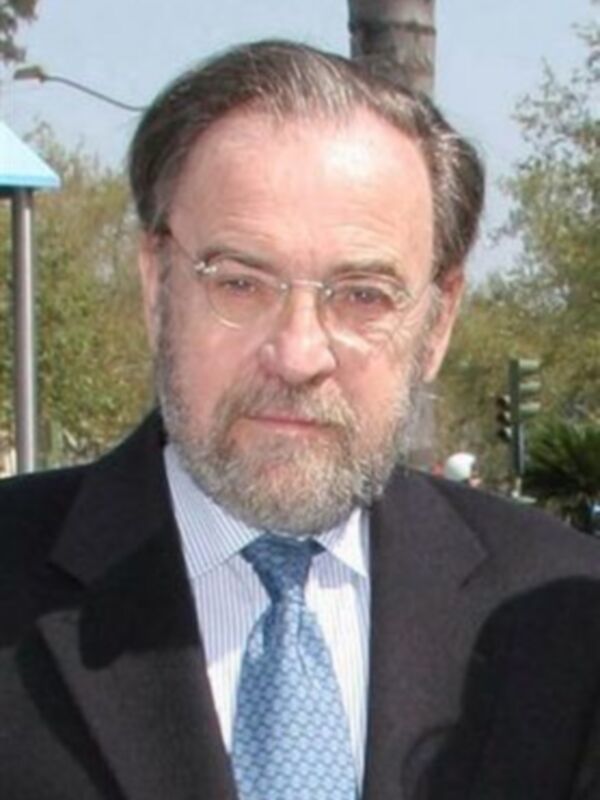 Un hombre con barba y bigote, llevando un traje negro, camisa azul claro y corbata. Está en una calle con árboles y un semáforo visible al fondo.