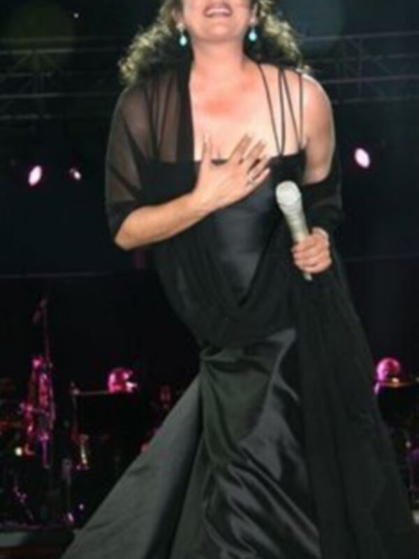 La cantante se encuentra en un escenario con luces de fondo, vestida con un elegante vestido negro que se mueve al ritmo de la música. La escena transmite una atmósfera de concierto o actuación musical.