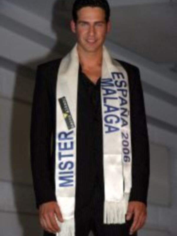 Un hombre vestido con un traje negro y una corbata blanca, sosteniendo una cinta que dice "Mister España Málaga 2006".