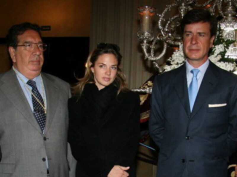 Tres personas en trajes formales posando juntas en un ambiente elegante con un candelabro de fondo.