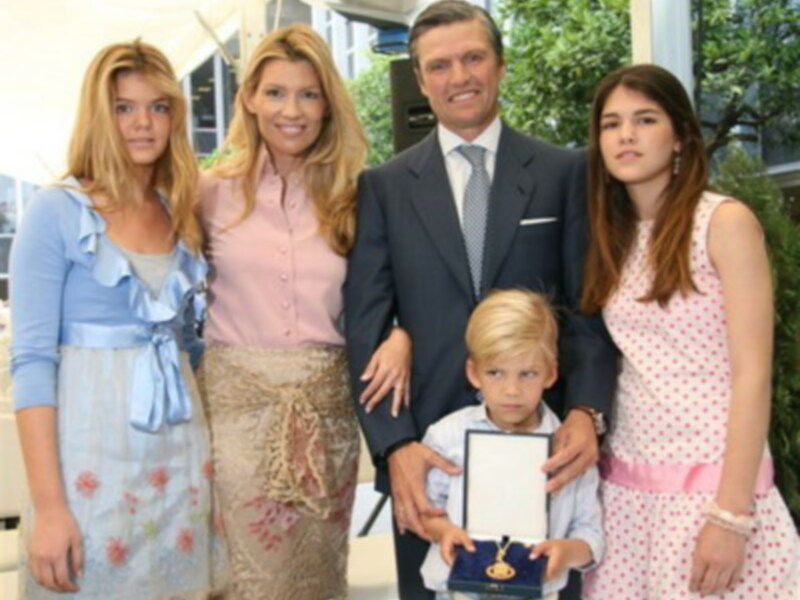 Una familia feliz con un padre y tres hijas posando para una foto. La madre lleva una falda beige con flores, la hija mayor tiene un vestido azul claro, el hijo menor sostiene un diploma y la hija más pequeña lleva un vestido rosa con lunares.