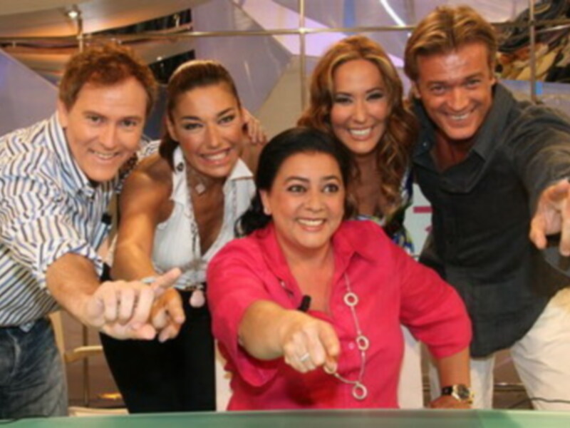 Un grupo de personas sonrientes y emocionadas en un estudio de televisión, posando con una mano extendida hacia la cámara.
