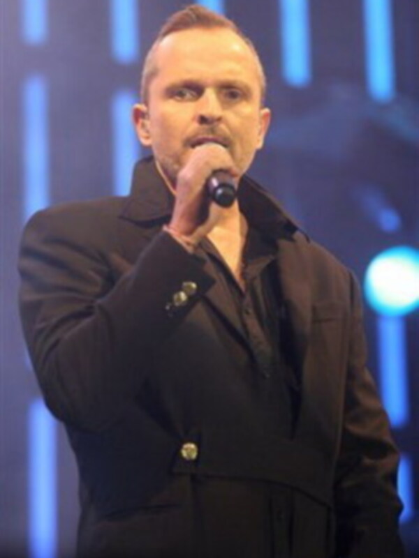 Un hombre con una chaqueta negra y un cuello alto, sostiene un micrófono en la mano izquierda. La iluminación es intensa con luces azules y blancas en el fondo, lo que sugiere un escenario de concierto o evento.