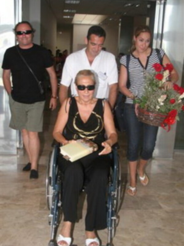 Una persona en silla de ruedas camina con una señora en brazos, mientras una mujer con un bolso y una florera camina detrás de ellos.