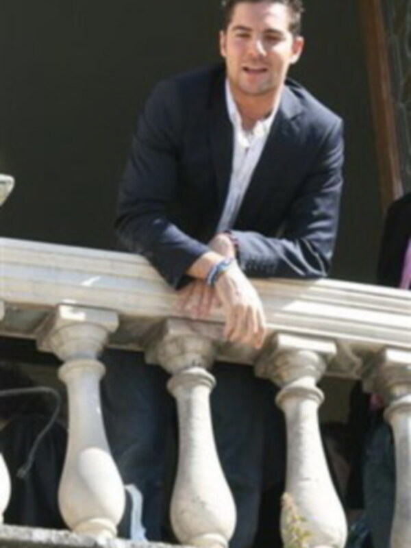 Un hombre con una camisa blanca y un chaleco negro está sonriendo desde el balcón de una casa.