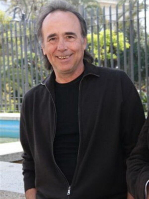 Un hombre con cabello gris y barba, vistiendo una chaqueta negra sobre un jersey negro. Fondo de una cerca metálica y vegetación.