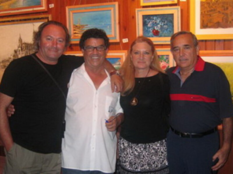 Cuatro personas sonríen frente a una pared de cuadros. La imagen muestra una reunión o evento en un ambiente cultural, posiblemente una galería de arte.