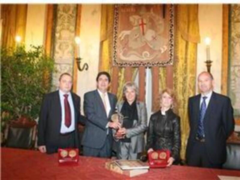 A cinco personas posando en una sala decorada con tapices y un escudo heráldico. La mesa frente a ellos tiene dos libros y una caja roja con un símbolo en el centro. La escena parece ser formal, posiblemente una ceremonia o reunión de negocios.