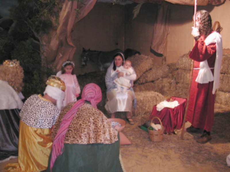 Escena navideña con personajes vestidos de época, posiblemente en un pesebre. Una mujer sostiene a un bebé mientras otras personas observan. La escena está ambientada en un entorno de paja y madera, con una figura vestida como ángel.